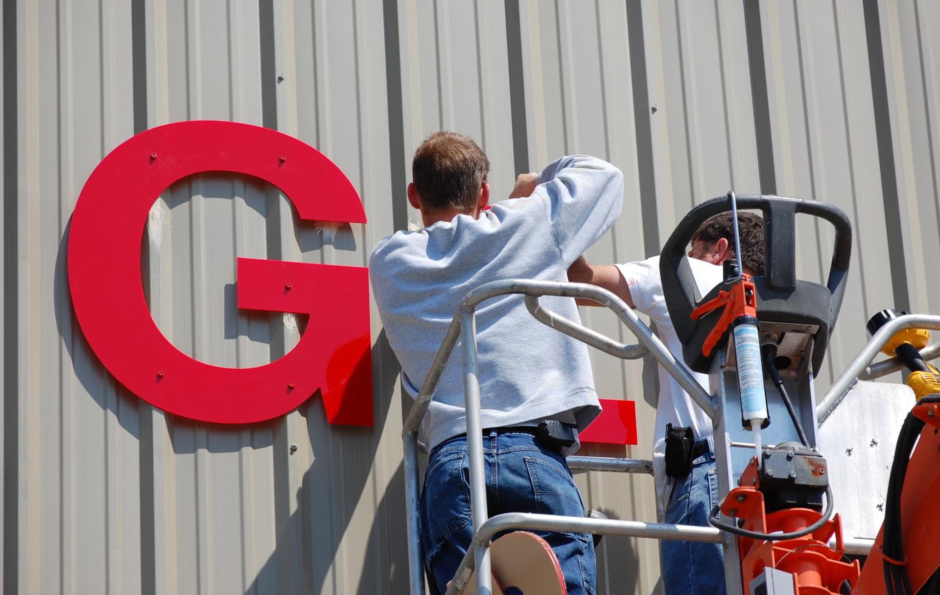 Two men are installing a large red letter g on the side of a building.