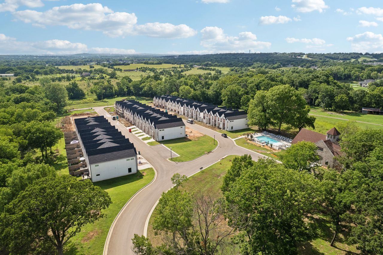 Aerial view of townhomes with black roofs, surrounded by trees, a pool, and green fields.