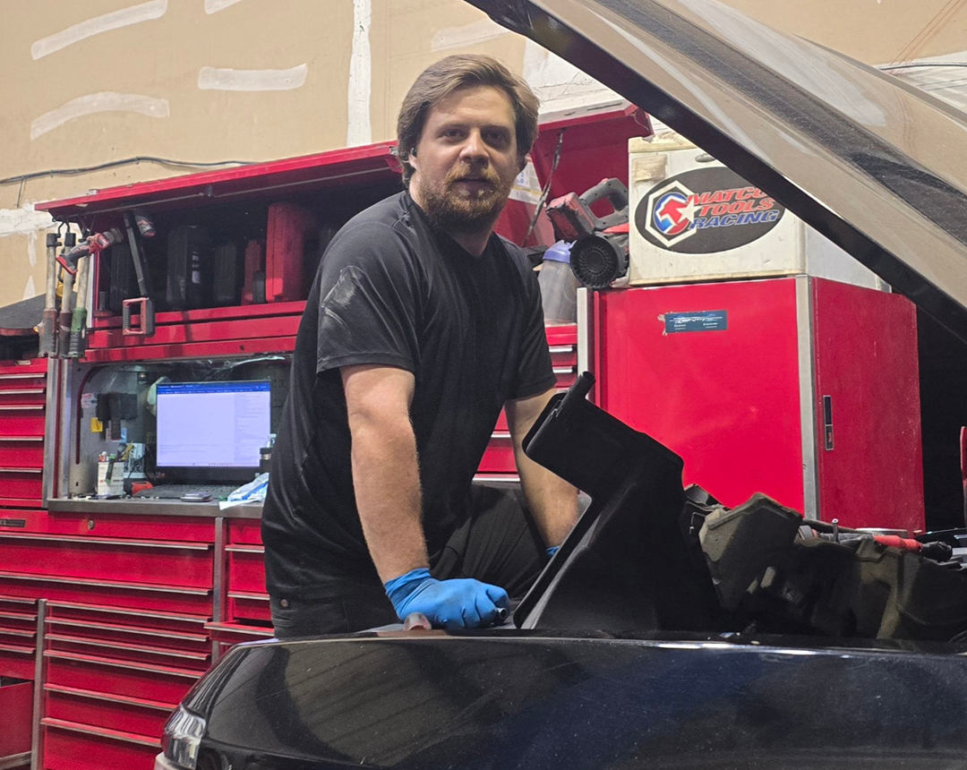Mechanic working on a car engine, wearing gloves. Garage setting, red tool cabinets, man smiling at camera. | Integrity Auto Repair
