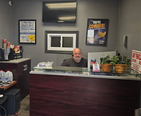 A man at a desk inside an auto shop. He has a gray beard and is wearing a gray shirt. | Integrity Auto Repair