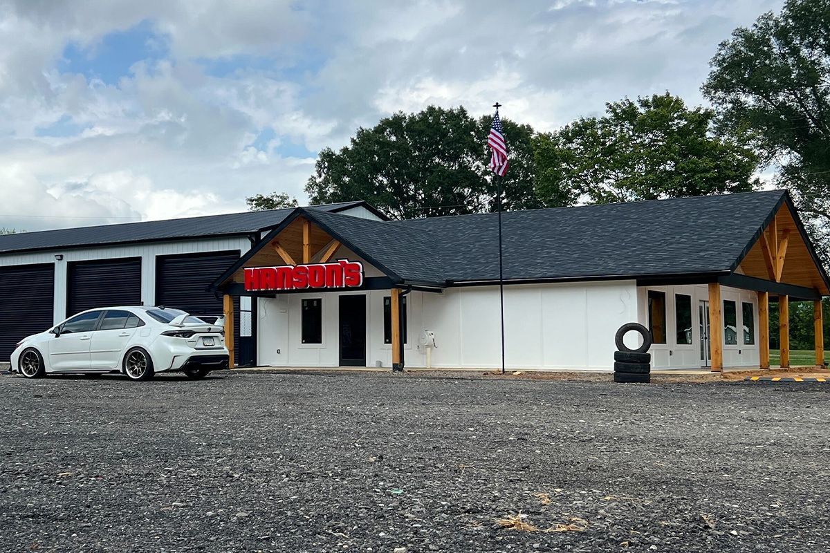 A white building with a black roof, storage units, and a white car parked outside on a gravel lot.