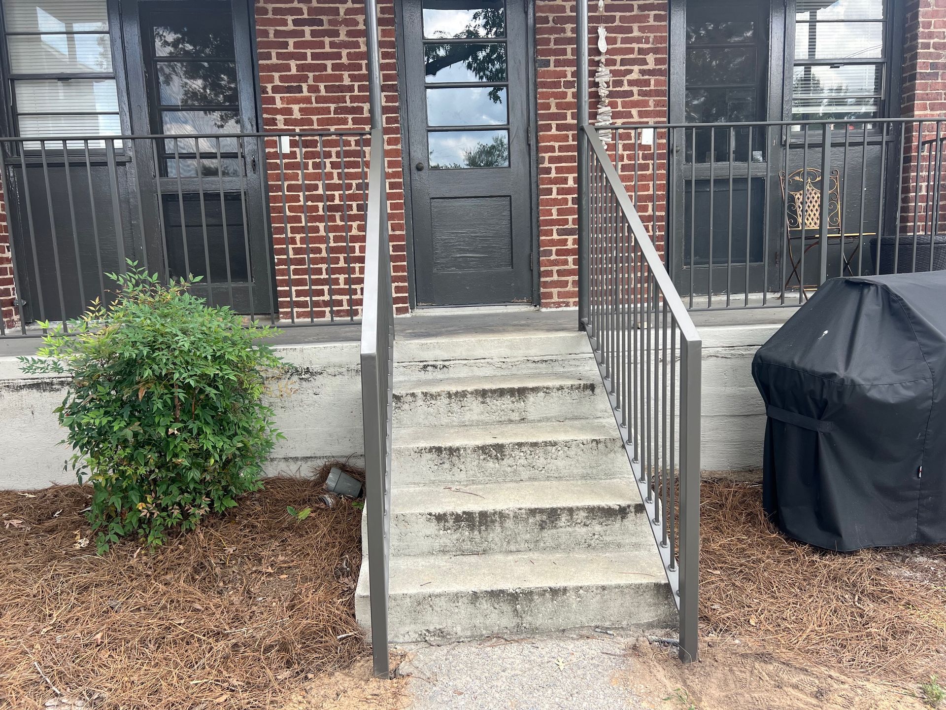 Exterior of building with steps, door, and metal railings. Brown mulch and small bush in front.