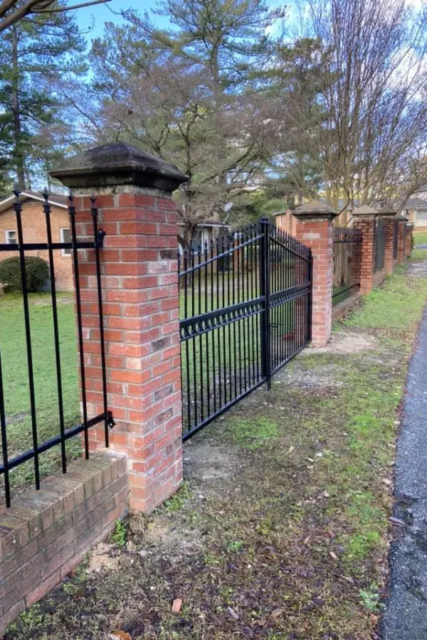 Brick and wrought iron fence with gate, grassy yard, trees in background.