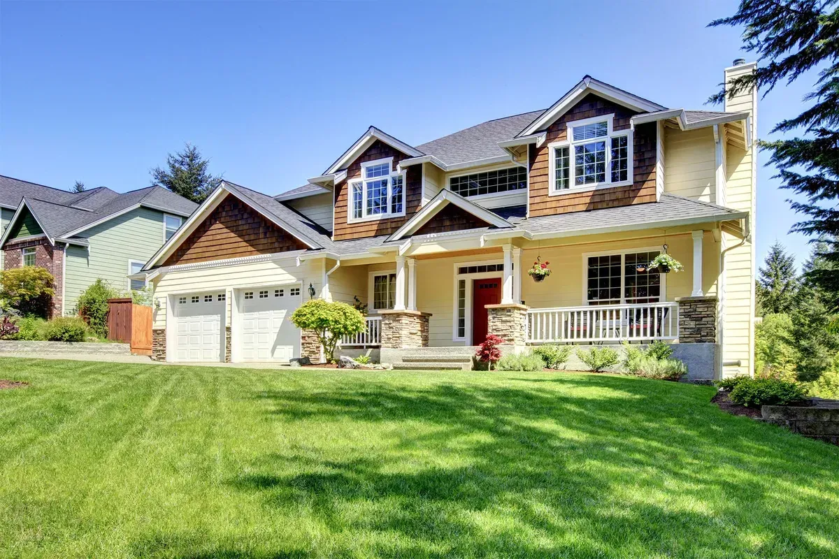 Two-story house with tan siding, brown roof, and green lawn on a sunny day.