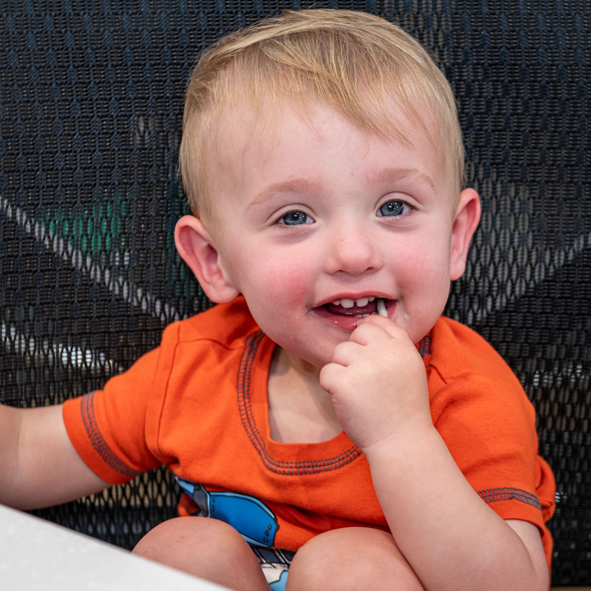 Smiling toddler with blonde hair and blue eyes, wearing an orange shirt, sitting and holding something to their mouth.