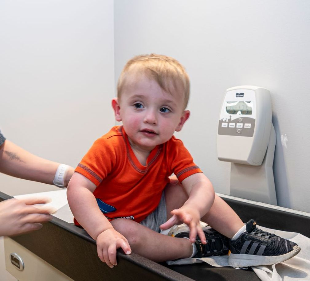 Child in orange shirt sitting on a medical exam table while a clinician assists nearby
