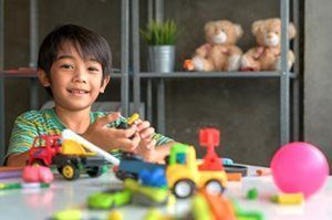 Child playing with toy cars at a table, with a pink ball and stuffed bears on shelves in the background.