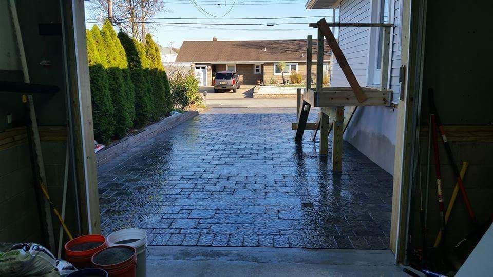 View from garage onto a brick driveway leading to a house, with green bushes and sky visible.
