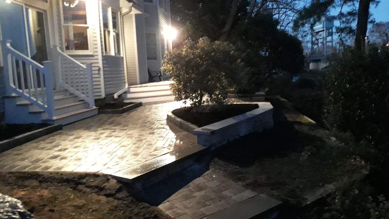 Night shot of a patio with a built-in planter and steps leading up to a white house with lights on.