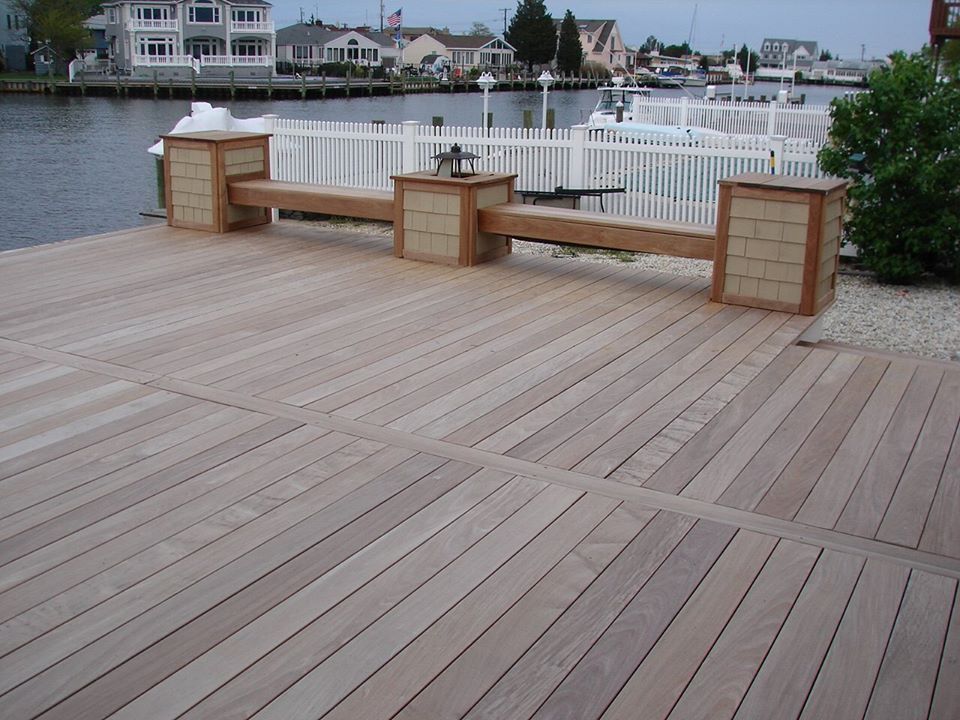 Wooden deck with built-in bench, waterfront view with white fence and houses in the background.