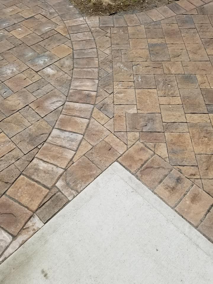 Stone paver walkway merging with a concrete sidewalk. Brown pavers form curves and straight lines.