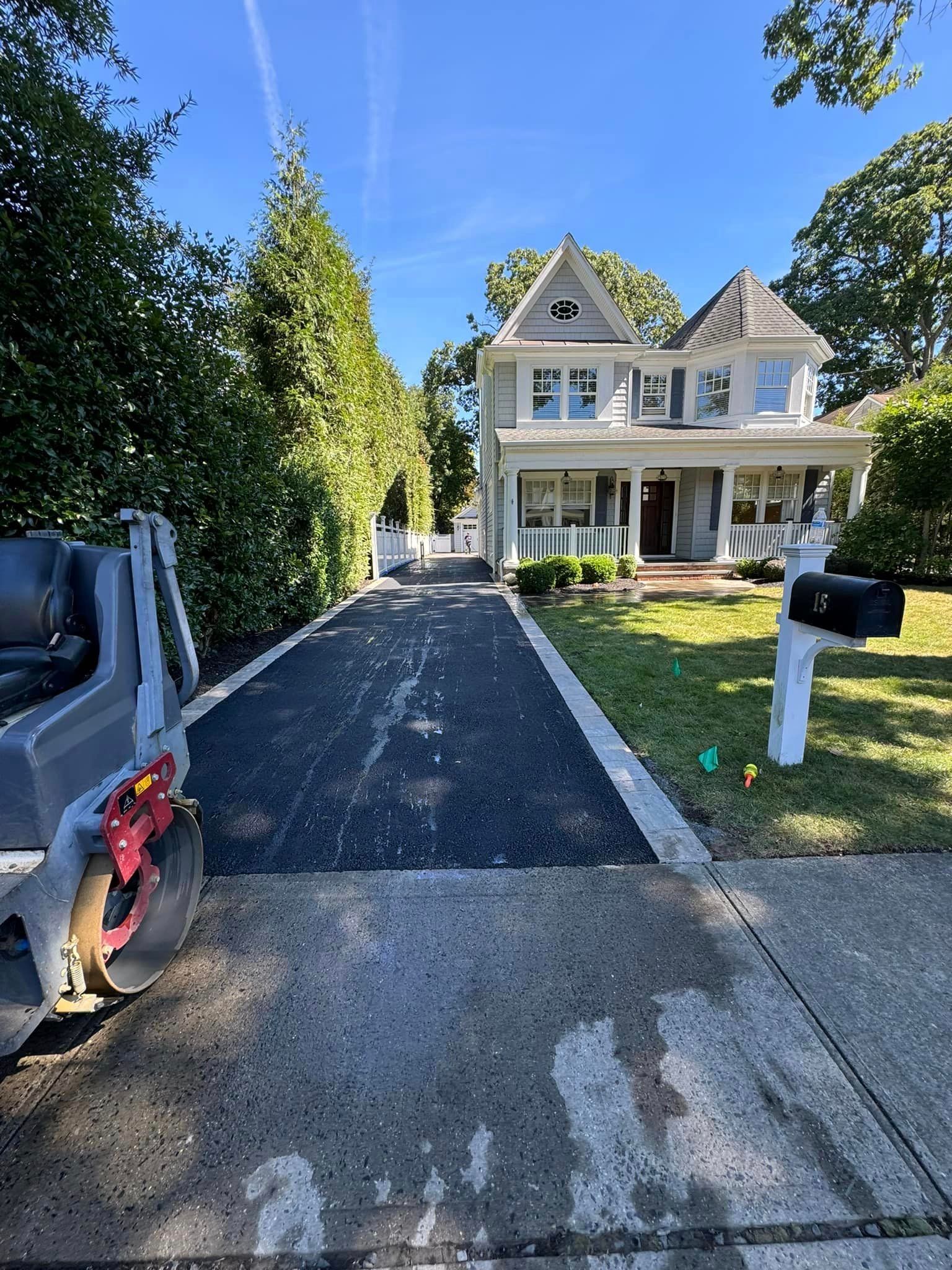 Newly paved asphalt driveway leading to a two-story house with a porch and green lawn.