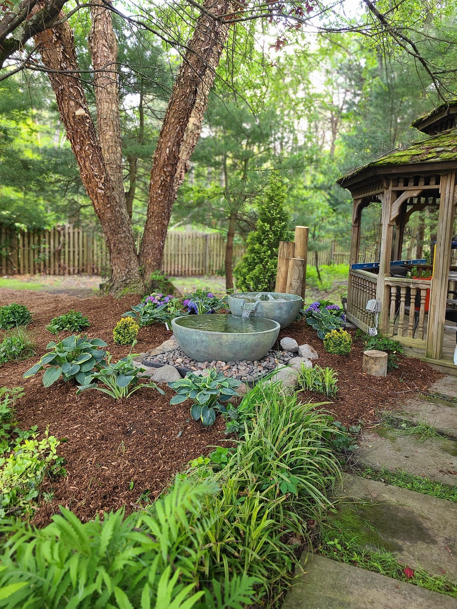 A garden with a water fountain, plants, and a gazebo near a forest.