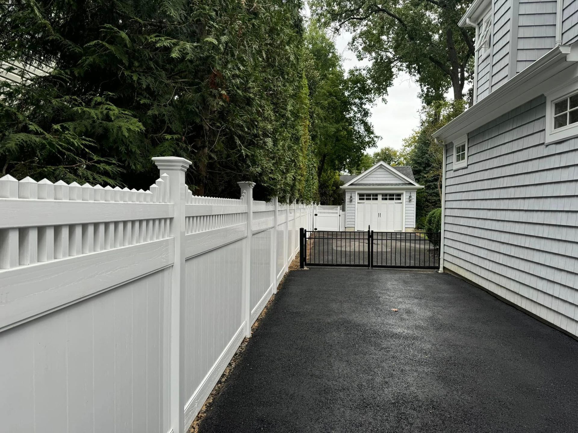 White fence along a black asphalt driveway leading to a garage. Gray house on right.