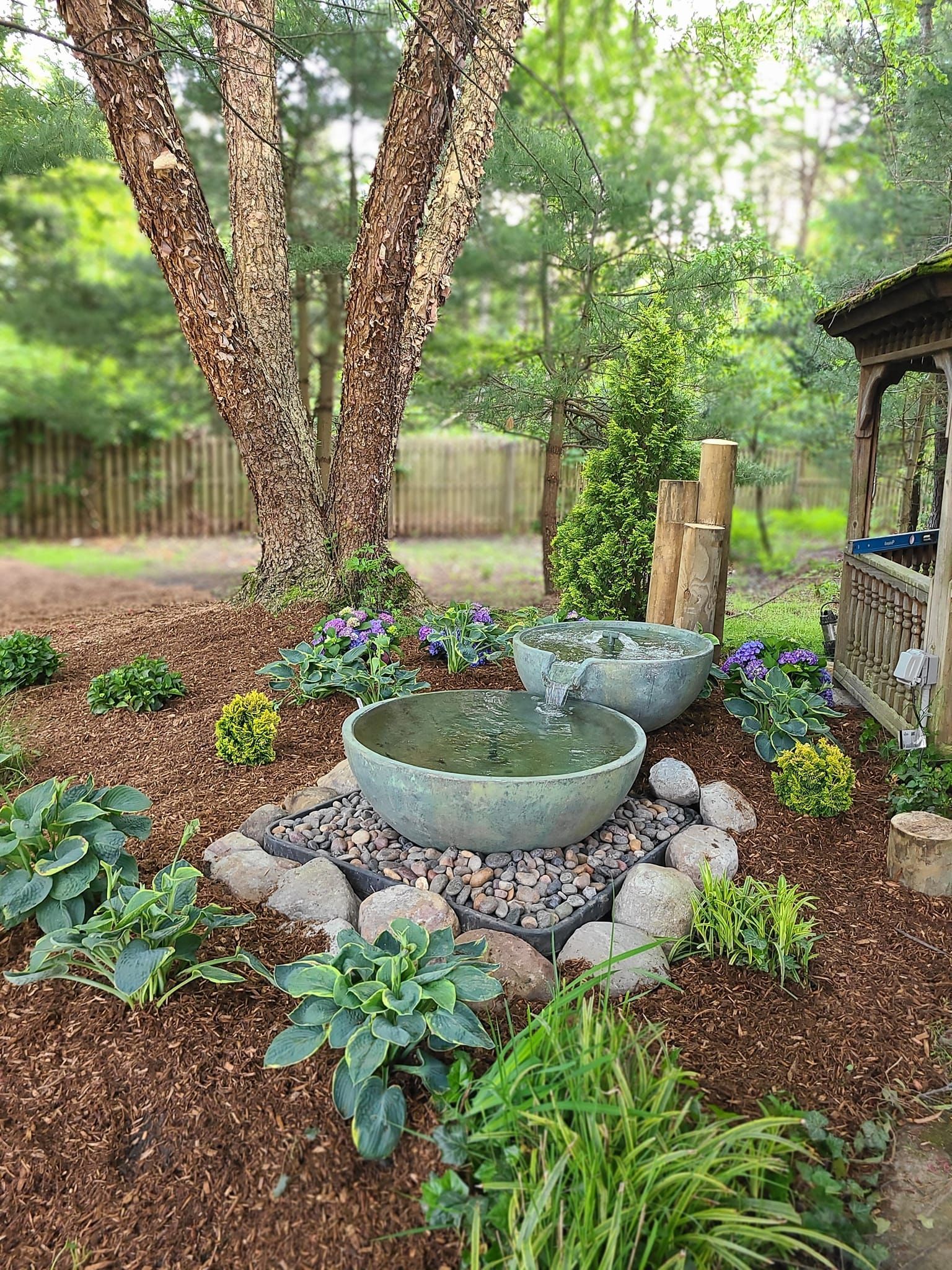 Water fountain in a garden with plants, wood chips, and a gazebo in the background.