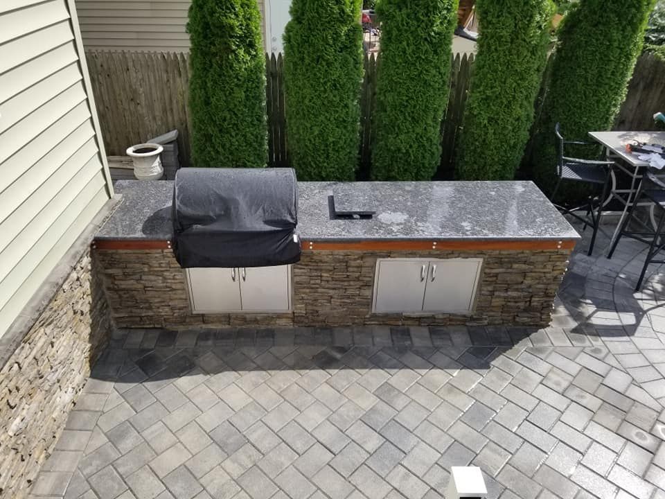 Outdoor kitchen with stone facade, stainless steel appliances, and dark countertop on a paver patio.