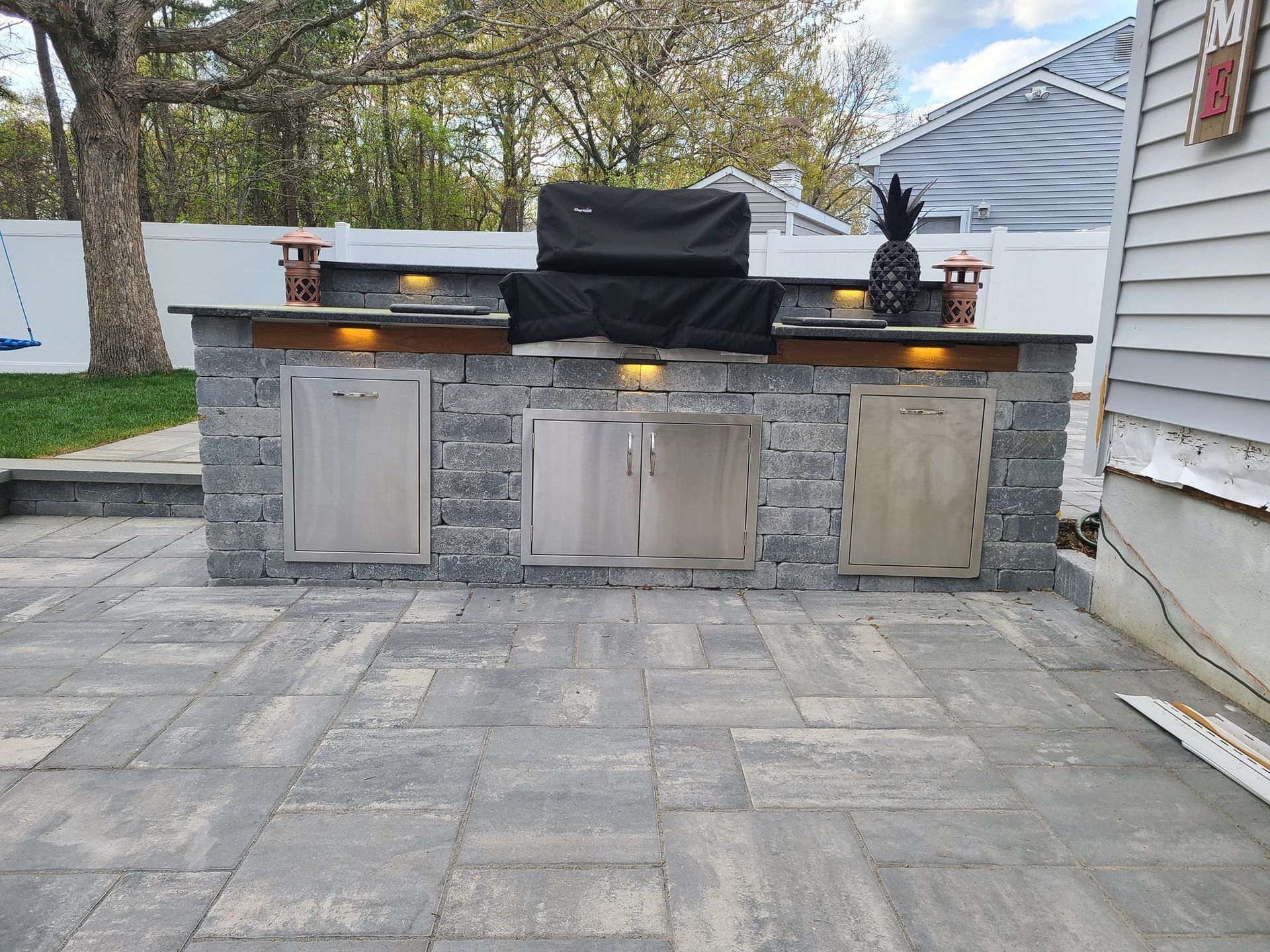 Outdoor kitchen with a grill, cabinets, and counter space, built into a block wall on a paved patio.