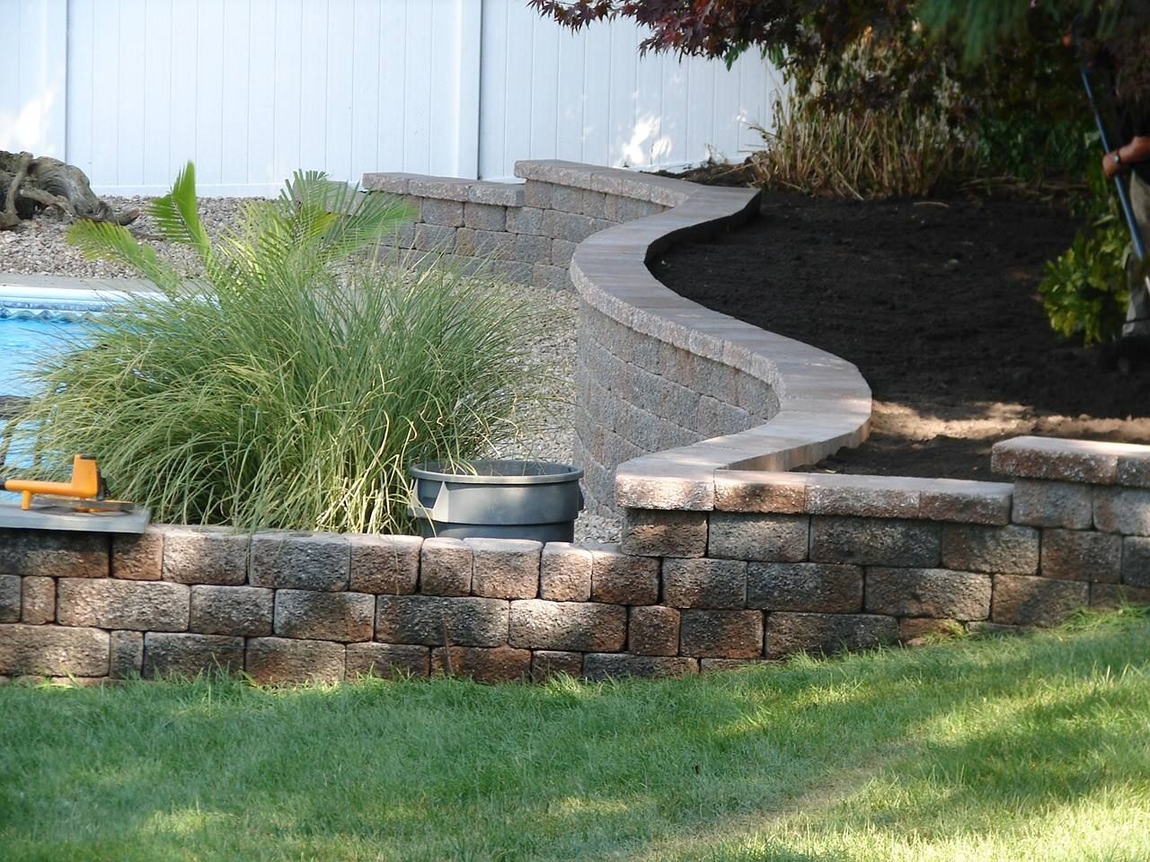 Retaining wall made of brown blocks, curving up a sloped yard with a pool in the background and green grass in the foreground.