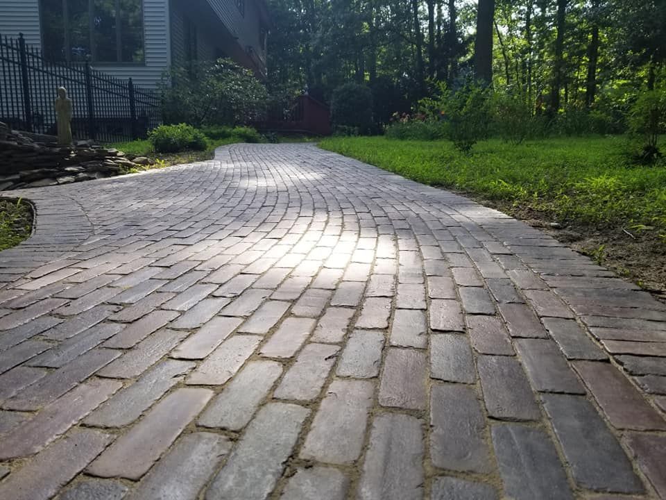 Brick driveway curves towards a house, with green grass and trees in the background. Sunlight gleams on the bricks.