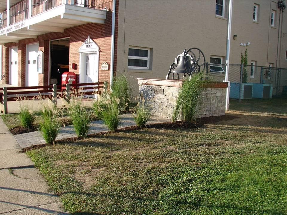 Exterior of a building with a bell monument on a stone base, green grass, and small plants.