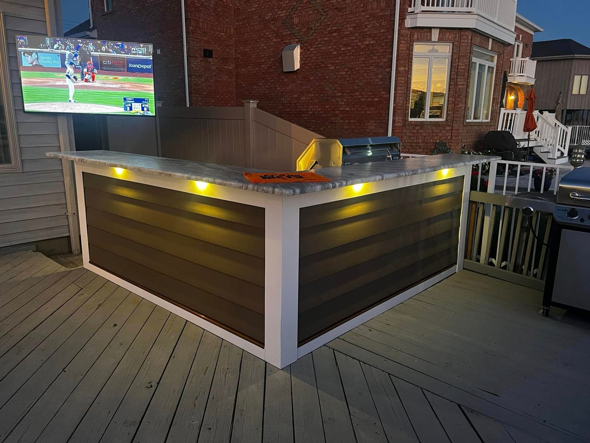 Outdoor kitchen with a TV, brown wood siding, granite countertop, and deck lighting.