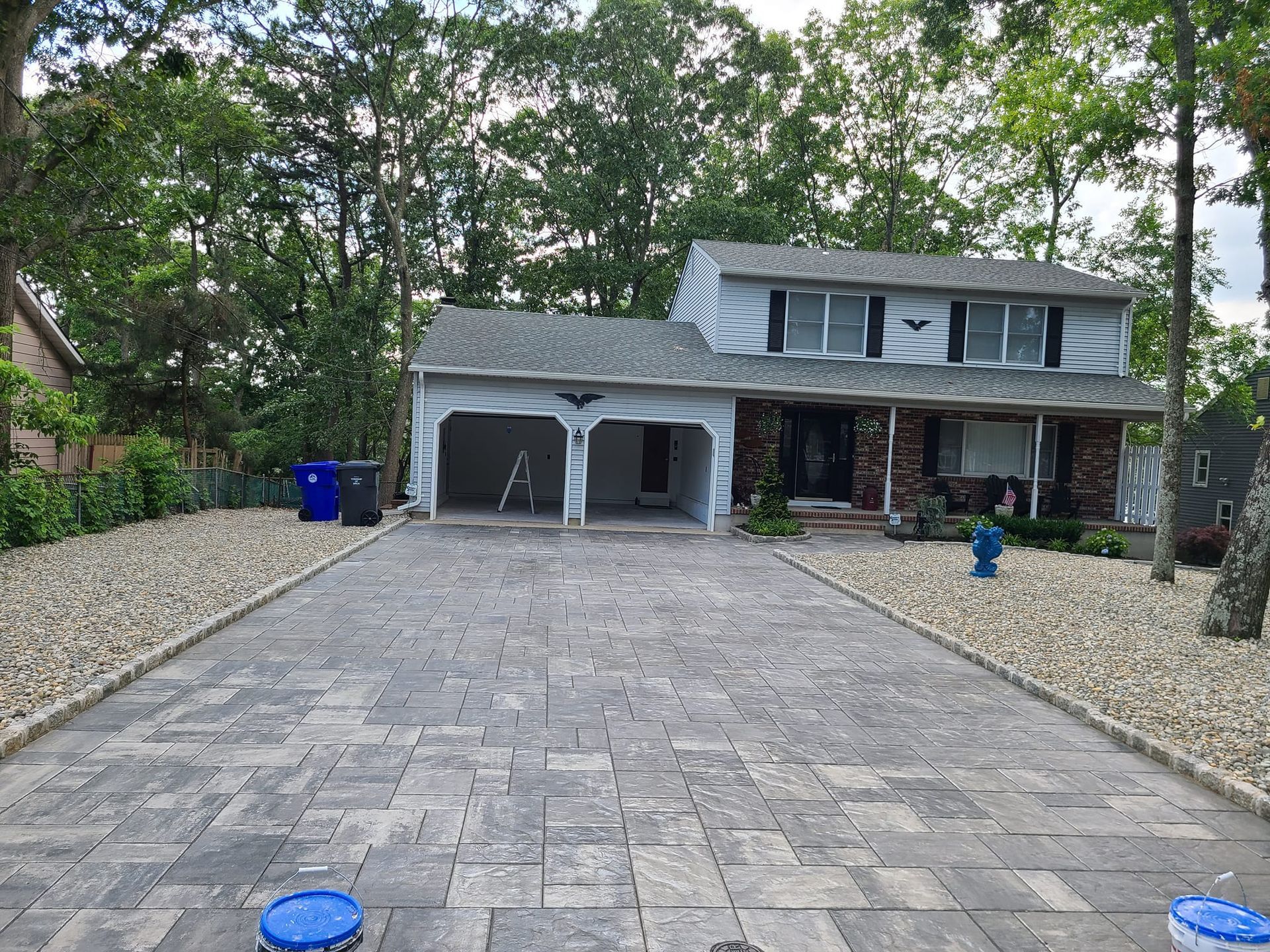 Brick paved driveway leading to a two-story house with a two-car garage. Beige gravel borders the driveway. Trees in background.