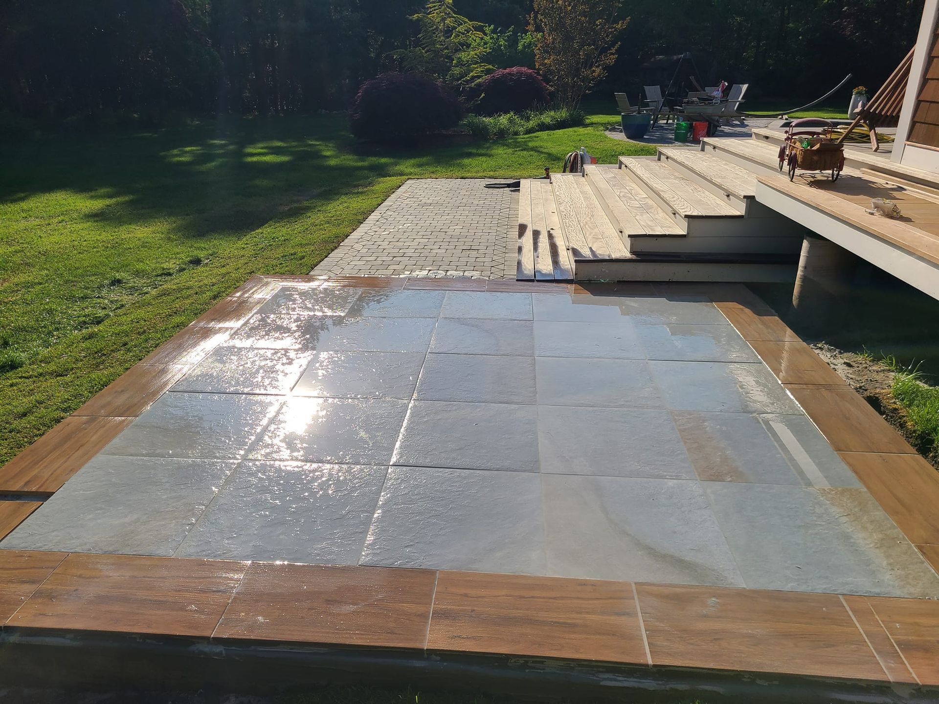 Wet blue stone patio with wood trim, near a wooden deck and green lawn in sunny backyard.