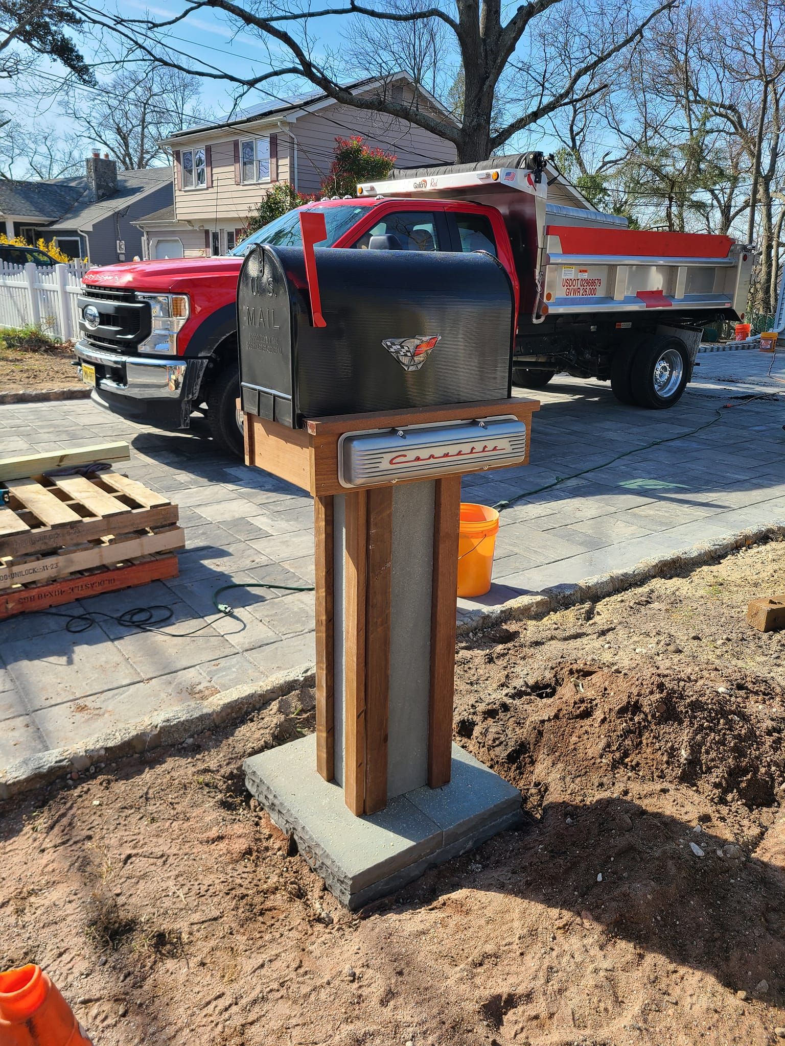 Mailbox on a wooden post with red truck in the background, near a sidewalk in front of houses.