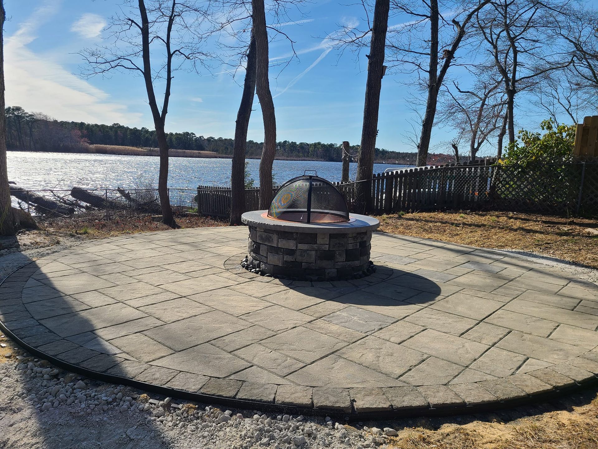 Stone fire pit on a circular stone patio by a lake. Bare trees and fence in background.