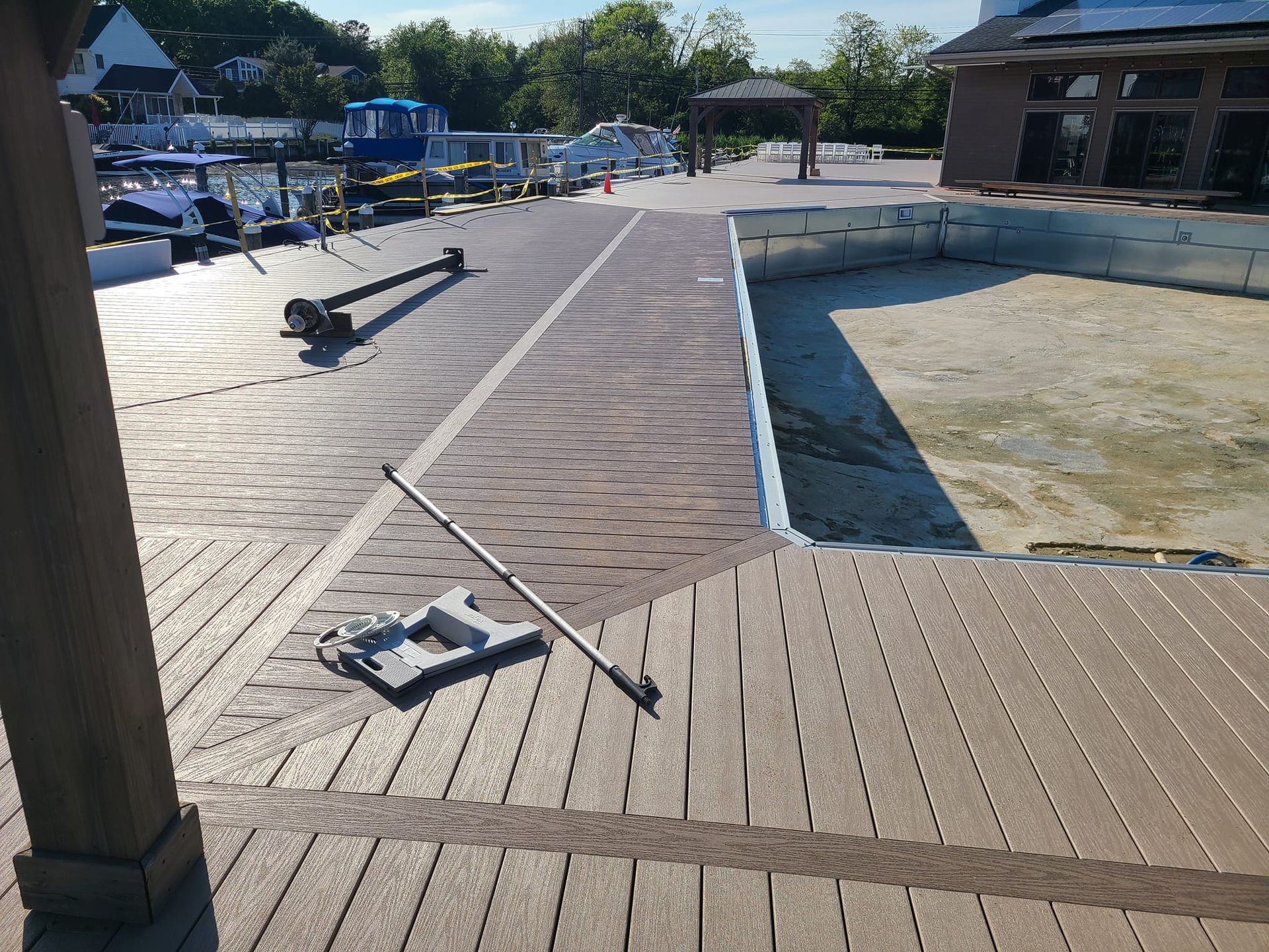Wooden dock with tools, adjacent to a textured pathway and a drained pool; boats in background.