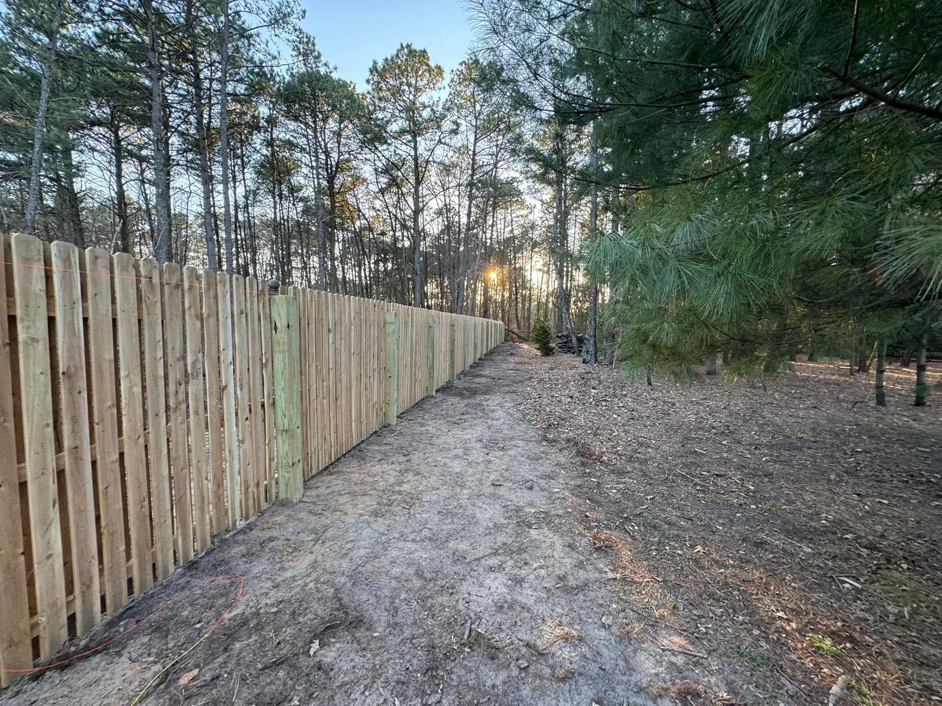 Wooden fence along a dirt path, with trees in the background and a bright sun.