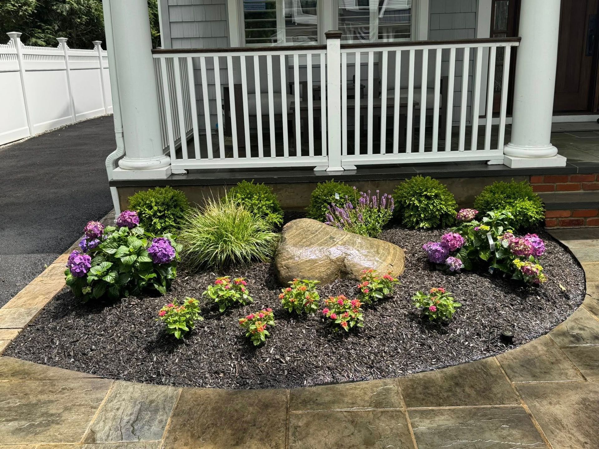Flowerbed in front of a house, with a large rock, various green plants, pink and purple flowers, and black mulch.