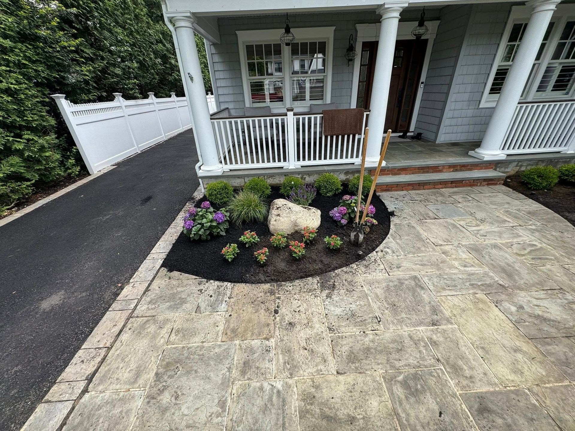 Stone walkway leading to a house with a porch. Landscaped flower bed with a large rock.