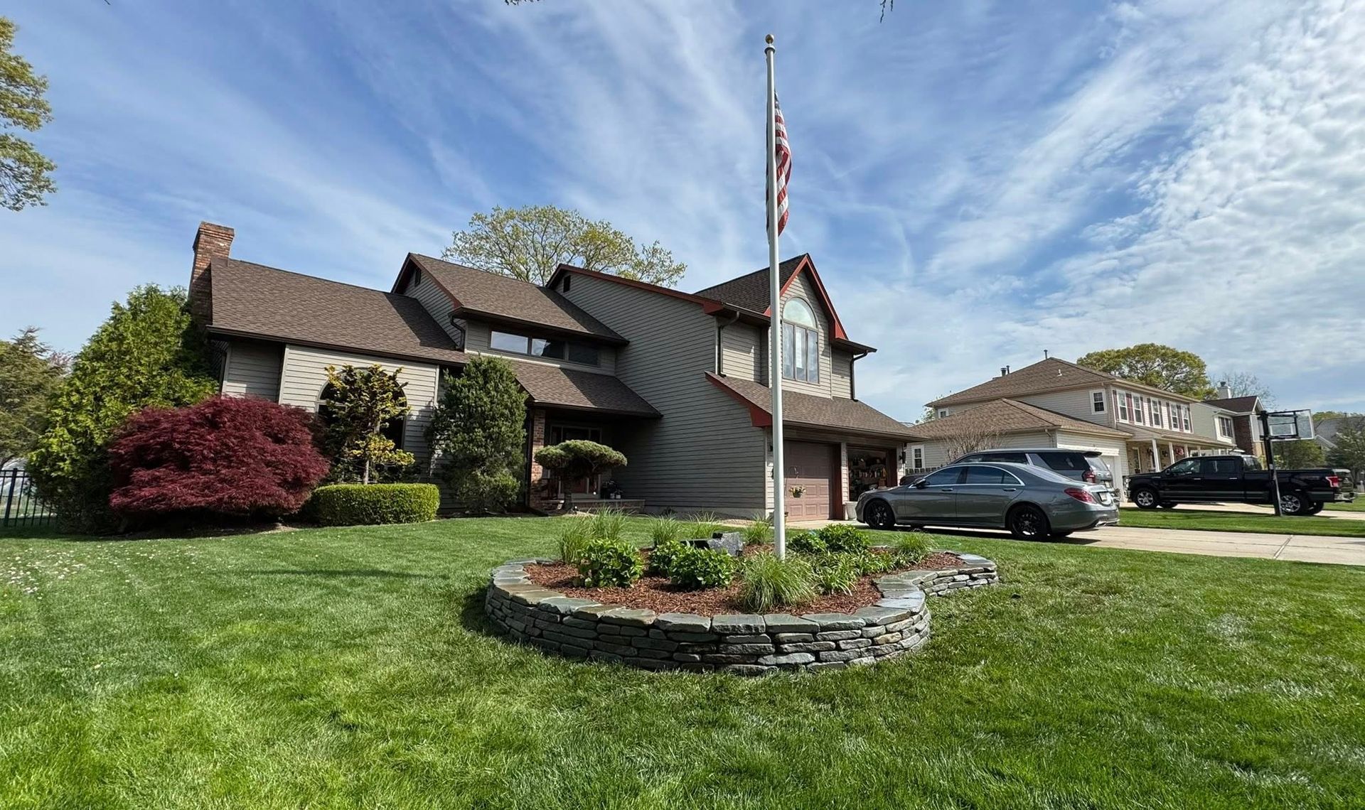 House with flag, garden bed, and cars on a sunny day. Brown roof, green grass, and blue sky.