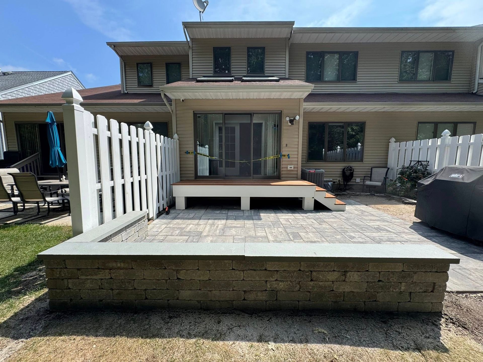 Backyard patio with raised stone wall, wooden deck, white picket fence, and a beige house.