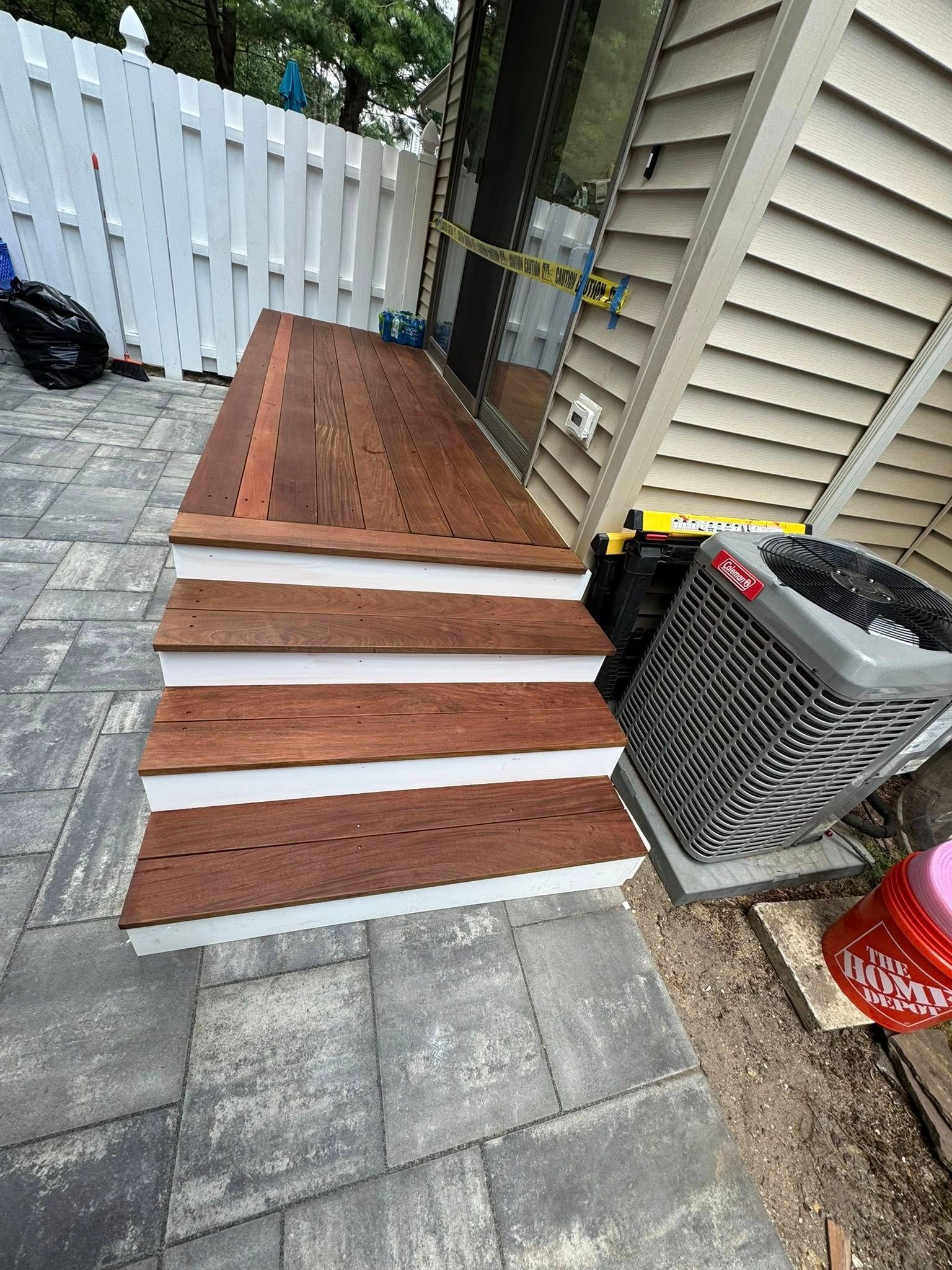 Wooden deck with steps, white trim. Next to a building and air conditioner, and a white fence in the background.