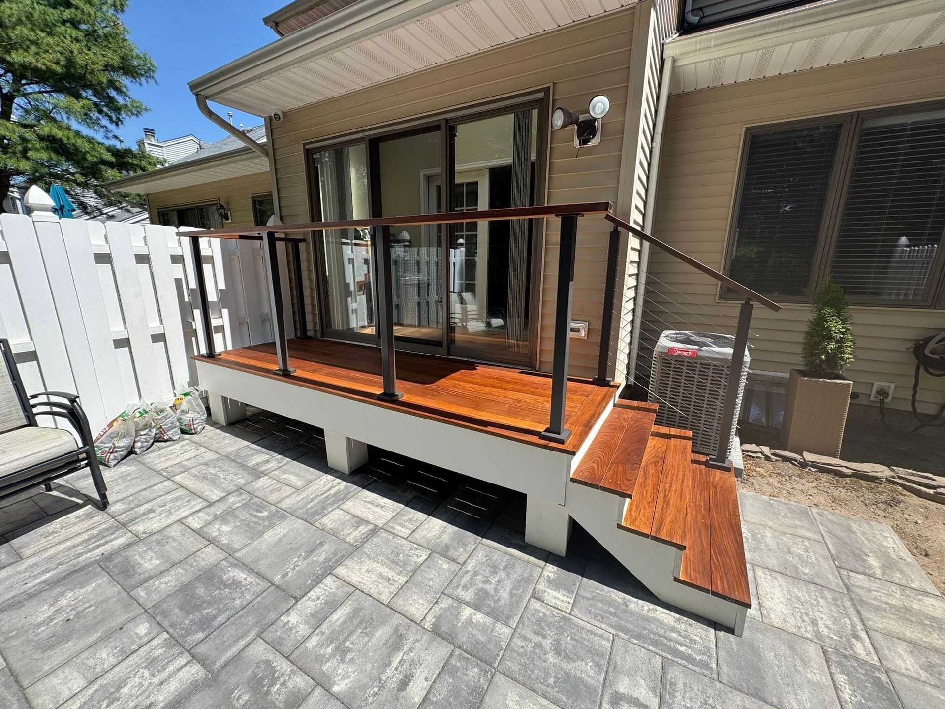 Wooden deck with steps, glass and dark railing, next to patio and white fence.