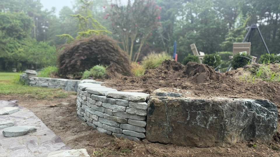 Stone retaining wall under construction in a garden with plants and soil.