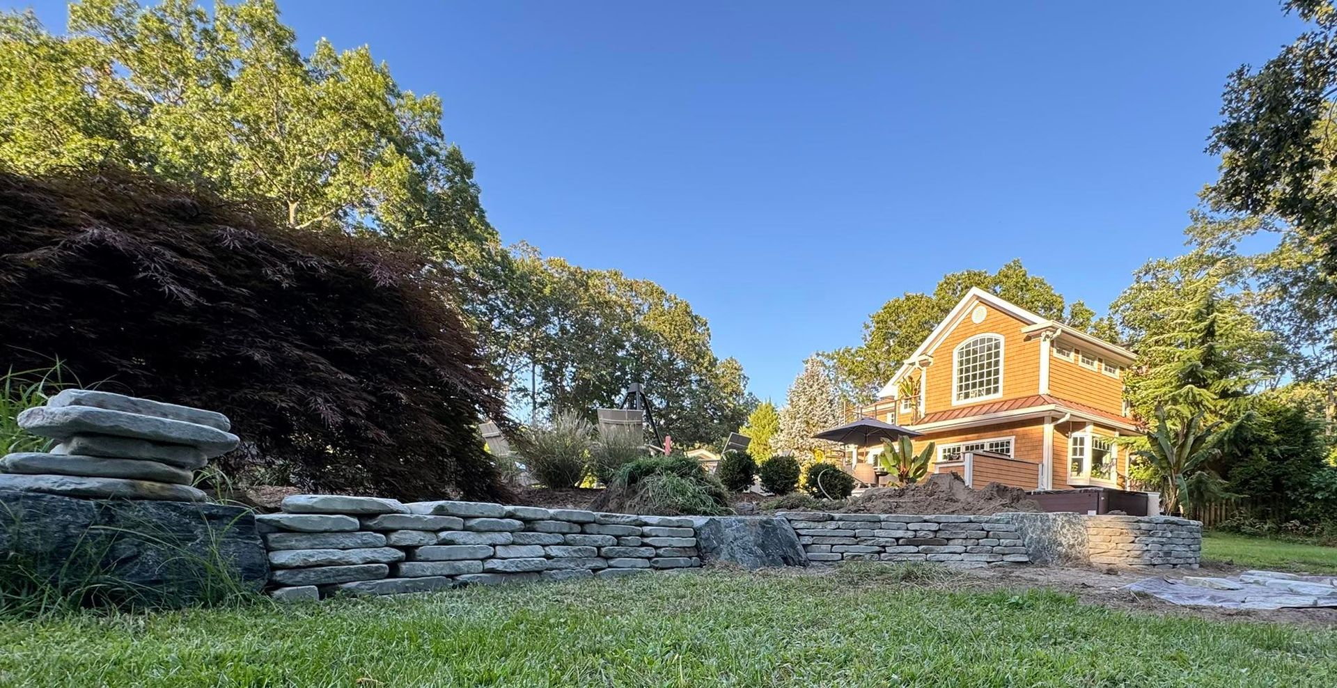 Orange house with arched window in sunny yard, stone wall in foreground, trees in background.