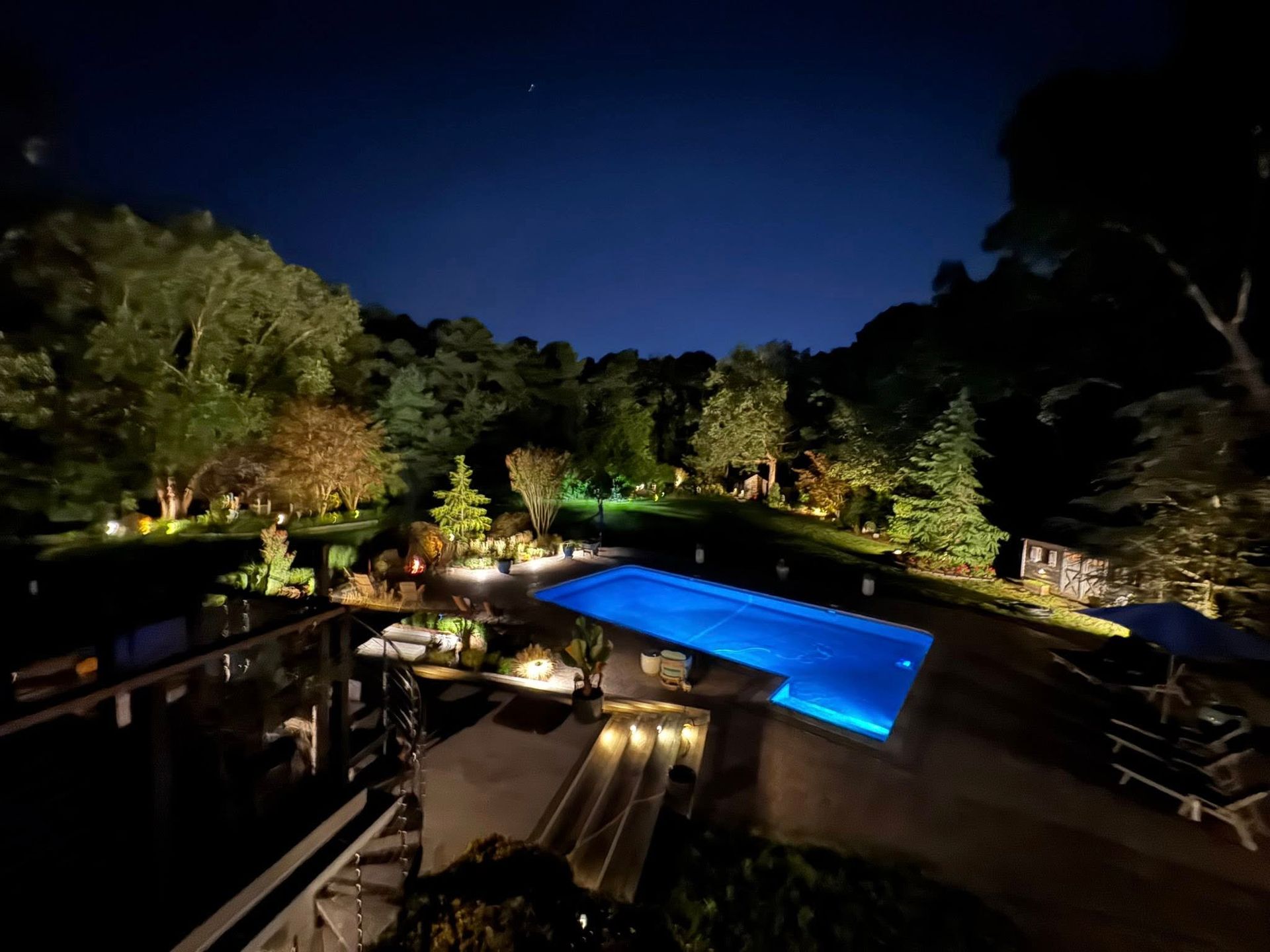 Nighttime view of a rectangular pool illuminated with blue light, surrounded by trees and landscaping under a dark blue sky.
