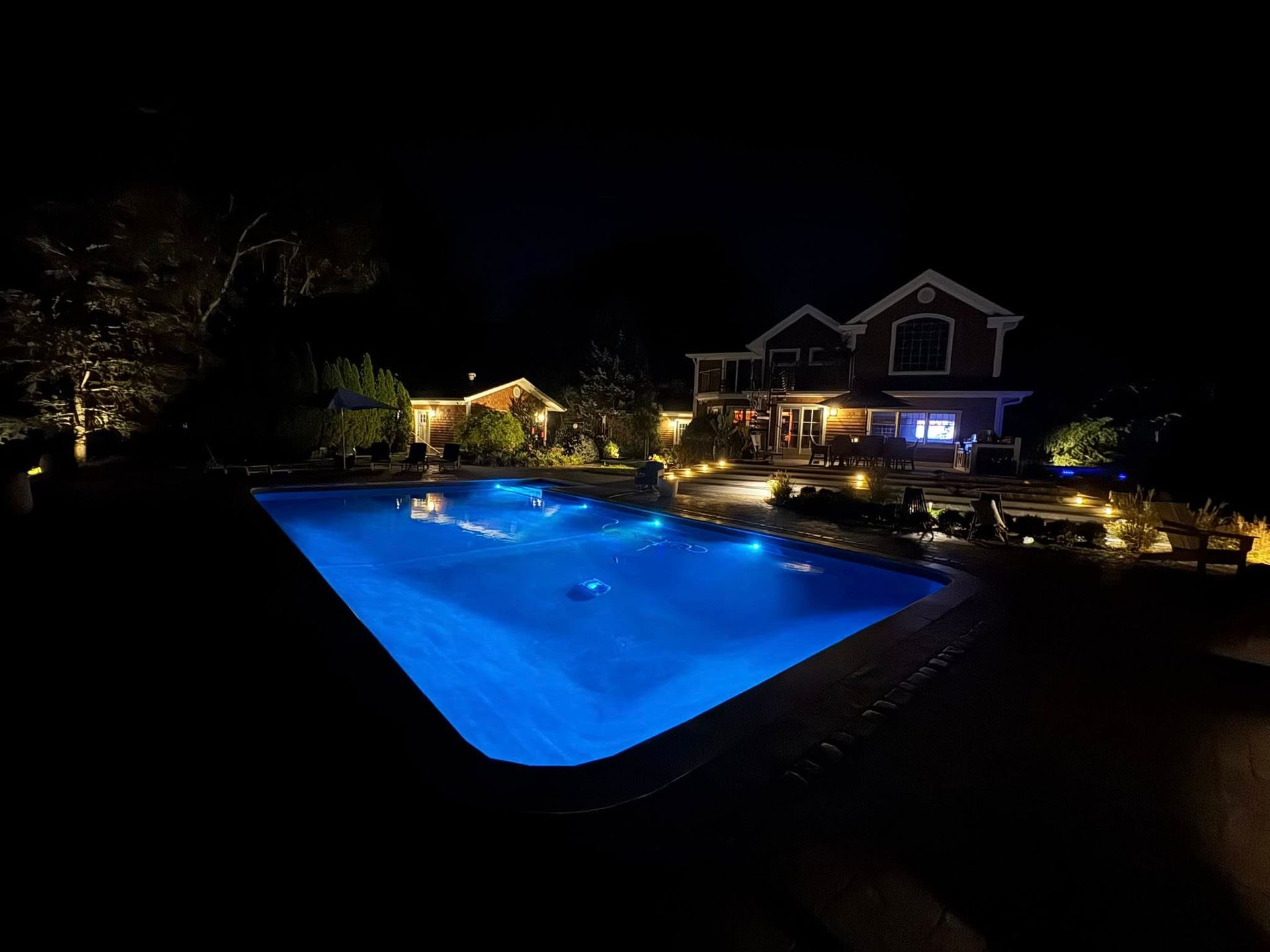 Nighttime view of a backyard pool illuminated with blue lights, near a two-story house with warm interior lighting.