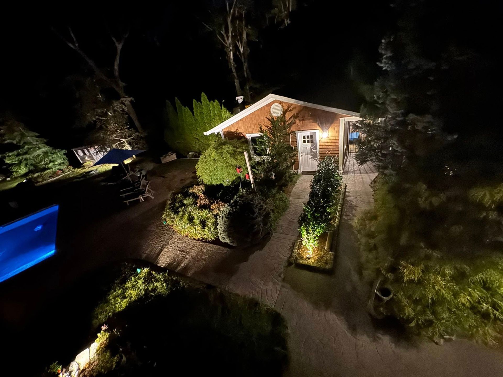 Small, illuminated cottage at night surrounded by greenery and lit pathways.