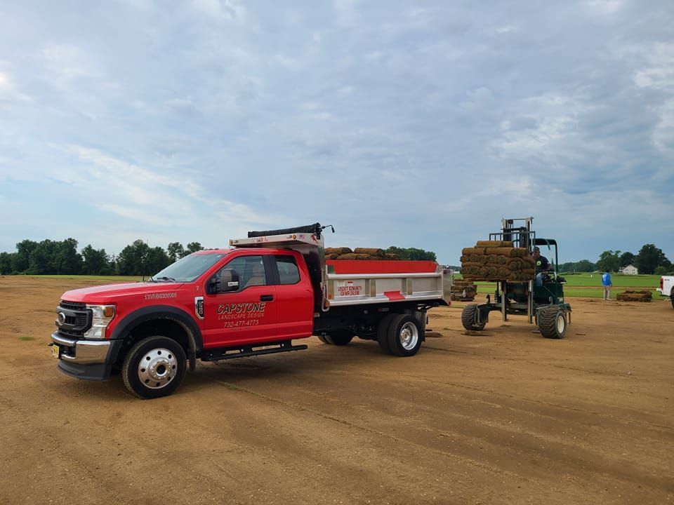 Red truck with a dump bed towing a tractor with a load of hay on a dirt field.