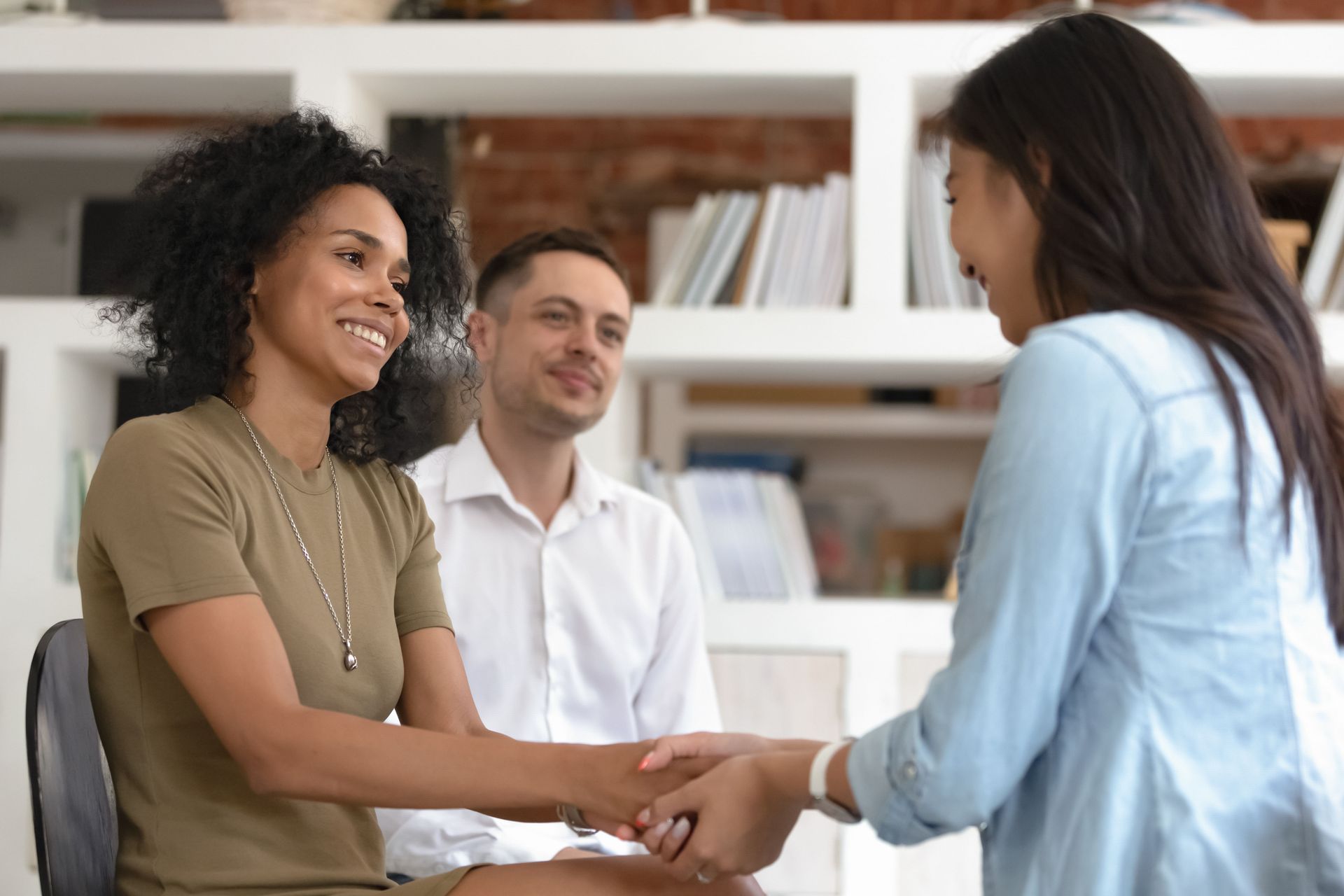 Woman shaking hands with another woman, while a man smiles in the background. Light-filled room.