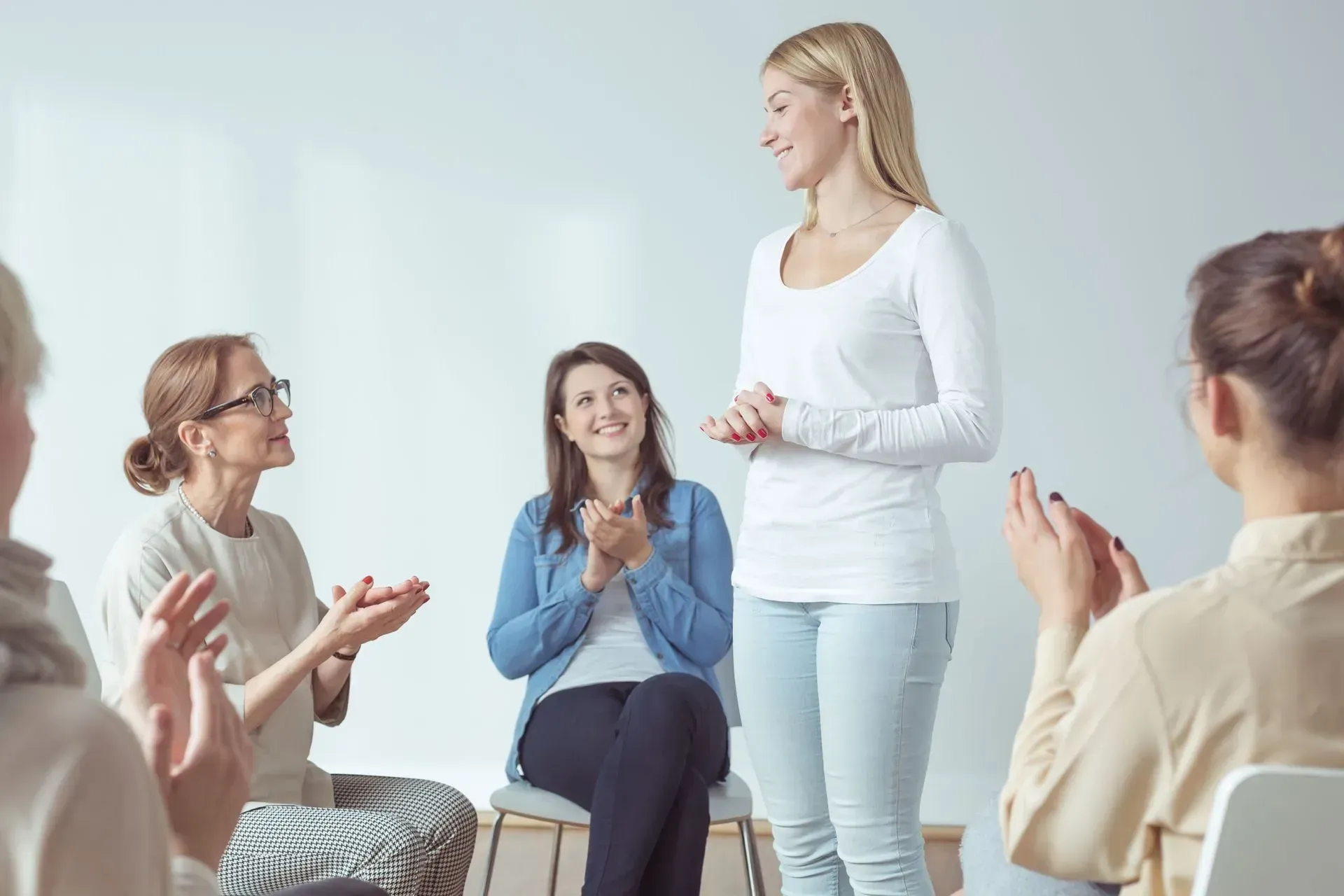 Woman stands, talking to group, all clapping in a circle, white room.