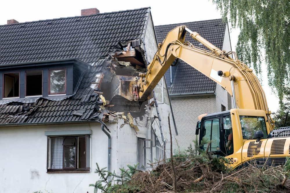 A Yellow Excavator Demolishing a White House With a Dark Gray Roof — Dirt Werx in Tathra, NSW