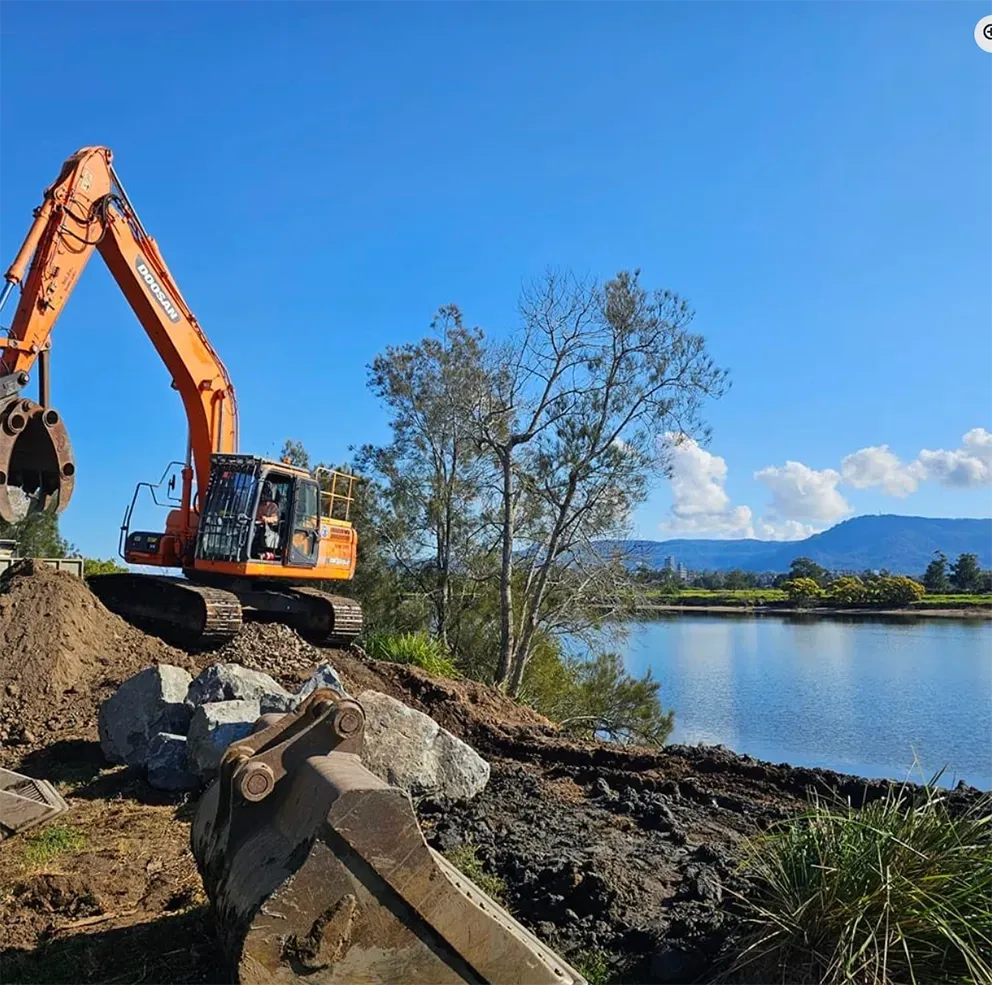 Orange excavator on a dirt pile near a river, blue sky, green trees — Dirt Werx in Narooma, NSW