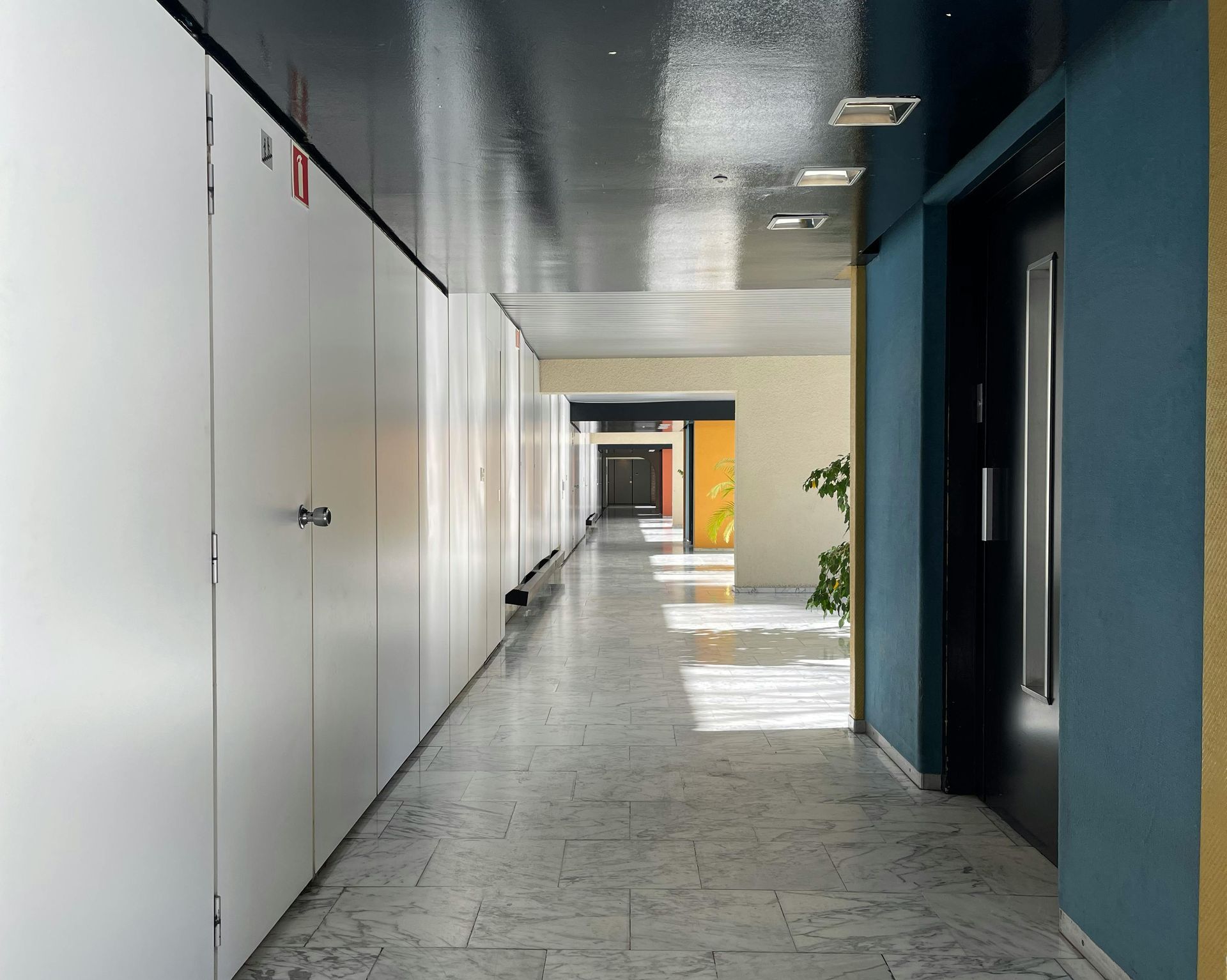 Long hallway with white lockers on the left, a blue wall on the right, and sunlight filtering through.