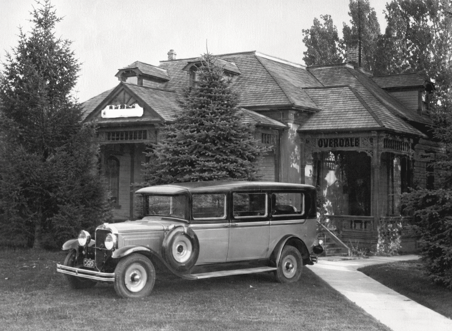 Vintage car parked outside a small stone building with a tiled roof and trees around it.