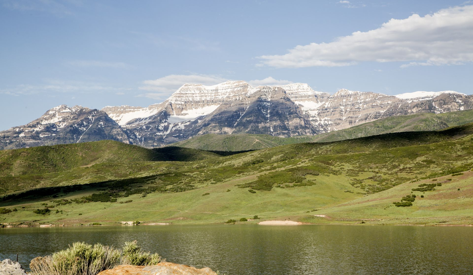Snowy mountains above green hills and a calm lake under a blue sky